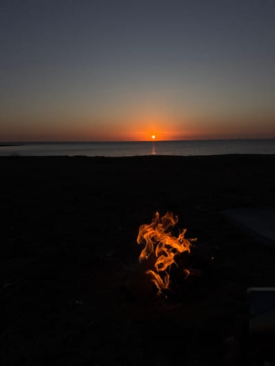 The image depicts a sunset over the sea, with a bonfire in the foreground. The sun is low on the horizon, casting an orange glow across the sky and reflecting on the water. The sea is calm, with a few distant lights visible on the horizon, possibly indicating a town or wind turbines. The foreground is dark and silhouetted, with the bonfire flames being the only source of light apart from the sunset. The bonfire appears to be in a natural setting, possibly a beach or a park, with dark ground visible around it. There are no people clearly visible in the image, though a hand holding a phone is partially visible on the right edge of the frame. The overall atmosphere is serene and peaceful, with a sense of warmth and tranquility emanating from the bonfire against the cool tones of the sunset and sea. The location is likely the coast of Thanet, United Kingdom, given the description of a sunset over the sea with potential offshore wind turbines.