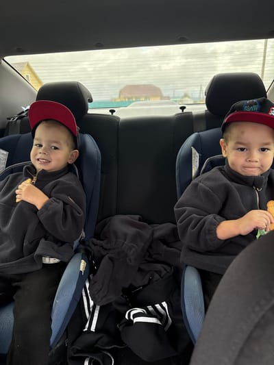 Two young boys are seated in car seats in the back seat of a vehicle, suggesting a trip to or from a location within Нижнеяркеево, Russia. The boy on the left, wearing a red baseball cap and a dark fleece jacket, is holding a cracker and smiling. The boy on the right, wearing a black baseball cap and a matching dark fleece jacket, is also holding a cracker. They are positioned side-by-side, facing forward. The interior of the car is visible, including the headrests of the front seats and the rear window, through which a cloudy sky and buildings of Нижнеяркеево can be seen. The car seats are dark blue, and there are black car seat covers and a dark blanket on the floor between the seats. The lighting suggests it is daytime.