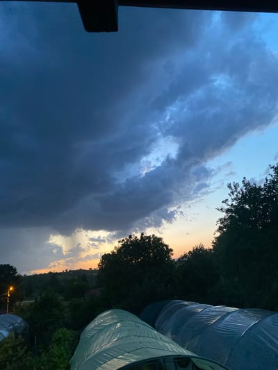The image depicts a cloudy sky at dusk, with dark storm clouds dominating the left side of the frame and lighter, sunset-tinged clouds visible towards the right. The horizon line is partially obscured by trees and possibly a distant hill, suggesting a rural or semi-rural location. A partially visible dark structure on the left side suggests an object in the foreground. On the right, there are light-colored rounded structures, possibly greenhouses, and the faint lights suggesting a settlement. It is not possible to confidently confirm the location as Konstantinovo, Bulgaria. There are no people visible in the image.
