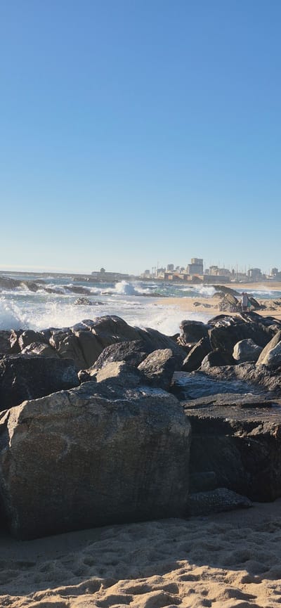 The image depicts a rocky shoreline with the ocean waves crashing against the rocks, creating white foam. The foreground is dominated by large, dark gray boulders and a sandy beach. In the mid-ground, the turbulent sea extends towards a distant coastline where a city skyline is visible. The city appears to be Vila do Conde, Portugal, with its characteristic buildings and a lighthouse. The sky is clear and blue, indicating a sunny day. A lone figure, possibly a person with a bag, is standing on the rocks in the mid-ground, looking out towards the sea. The scene captures a natural coastal environment with urban elements in the distance, likely taken from the area of the Praia do Forno or the mouth of the Ave River.