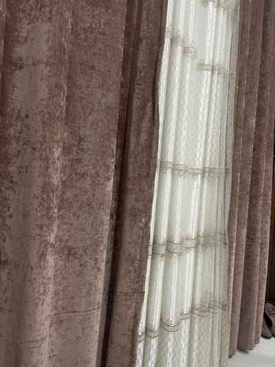 The image displays multiple rolls of fabric or curtains, arranged horizontally and viewed from a slightly elevated perspective looking downwards. The scene primarily showcases various textile textures and patterns.

In the upper portion of the image, several rolls of a textured, muted brown fabric are stacked. This material appears heavier and has a somewhat mottled or brushed texture. In the top right corner, a pair of light-colored, possibly beige or grey, slippers or soft shoes is partially visible, resting on top of one of these rolls, suggesting a domestic or personal storage setting.

Below these initial brown rolls lies a prominent section of lighter-colored fabric, which appears to be a sheer or semi-sheer material, likely intended for curtains or drapes. This fabric is rolled into distinct horizontal cylinders, with a base color of off-white or cream. It features a repeating decorative pattern: there are approximately five to six visible thicker vertical bands that are embroidered or woven with a light gold or silver thread, showcasing a delicate floral or leaf-like motif. Between these more ornate vertical bands, the sheer fabric displays a finer, subtle diamond-grid pattern, also in a shimmering thread.

Beneath the sheer, patterned fabric, a large section of a darker brown or taupe fabric is visible. This material appears significantly thicker and heavier than the sheer fabric, suggesting it might be a blackout or main curtain fabric. Its texture is notably rich and uneven, resembling distressed velvet or a similar plush material with a mottled, almost worn appearance, exhibiting variations in lighter and darker tones across its surface. Faint vertical creases or seams are visible within this lower brown fabric, indicating folds or how it might be constructed when hung.

The overall impression is one of textiles, possibly for interior decoration or window treatments, highlighting different fabric types and designs in a stacked arrangement.