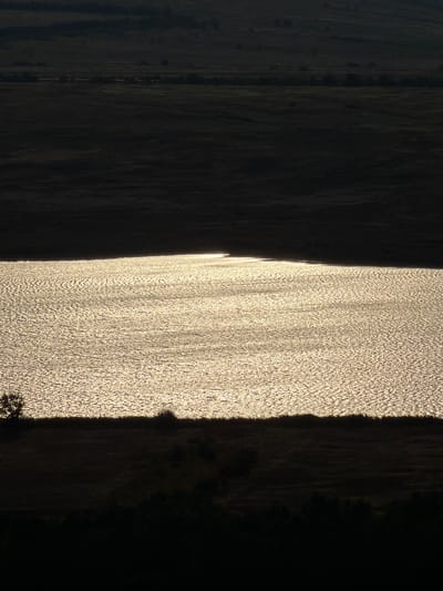 The image depicts a wide, tranquil body of water, likely a lake or a slow-moving river, shimmering with sunlight. The water's surface is rippled, creating a textured pattern of light and shadow. The scene is dominated by dramatic contrasts between light and dark. A dark, silhouetted landmass, possibly hills or mountains, forms the background, stretching across the upper portion of the image. The foreground also consists of dark, silhouetted land, featuring the faint outlines of vegetation, including what appears to be a lone tree on the left side and low-lying bushes. The overall atmosphere is serene and somewhat mysterious due to the strong backlighting and dark silhouettes. The specific location in Gornyi Balyklei, Russia, is not visually identifiable from the image itself; the scene is characteristic of a natural landscape.