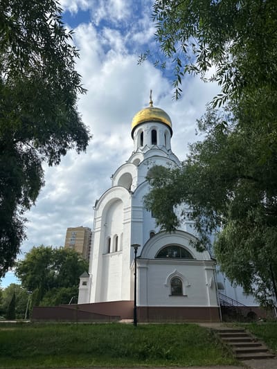 This is a vertical, low-angle, outdoor photograph of a white church with a golden dome. The church has arched windows and decorative architectural details. Lush green trees with dense foliage frame the top and bottom of the image, creating a sense of being under a canopy. Sunlight filters through the leaves, dappling the scene. The sky above is a mix of blue and white clouds, suggesting it is daytime, likely late morning or early afternoon, with partly cloudy weather.
In the background, to the right of the church, a tall, multi-story residential building with numerous windows is visible, indicating an urban or suburban setting. A patch of manicured green grass and a paved pathway run alongside the church. A lamppost stands on the right side of the pathway. The overall impression is one of peace and serenity, with the natural elements of trees and sky complementing the spiritual architecture. There is no visible text in the image. The scene is set in Kotelniki, Russia.