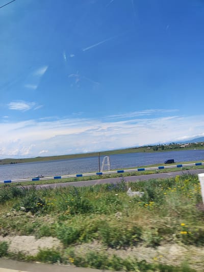 The image shows a wide, still body of water, likely a reservoir or lake, with a clear blue sky and scattered white clouds above. The water reflects the sky, creating a sense of depth. In the foreground, there is a green, grassy embankment with some yellow wildflowers. A road runs parallel to the water's edge, separated by a low barrier with blue and white markings.

On the far side of the road, by the water, a dark-colored van is parked. Further in the distance, across the water, a small village or settlement with houses is visible on a gentle slope, backed by rolling hills and mountains. The overall impression is one of a serene, rural landscape on a sunny day.

The presence of a van and the road suggests human activity, but there are no distinct individuals clearly visible in the image. The environment is natural, with a focus on the landscape and the water body. The scene is static and peaceful.
