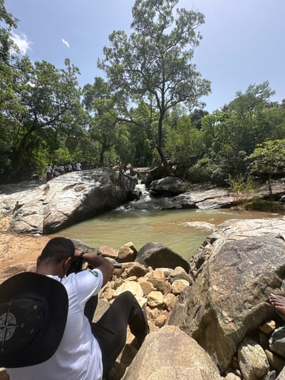 A medium shot captures a person from behind, seated on large rocks by a river in a natural setting, possibly Fobur, Nigeria. The individual is wearing a white t-shirt with a green logo and black pants, accessorized with a black hat featuring a compass design. They are holding a camera, seemingly taking a photograph of the scene.
The environment is lush and green, with dense trees forming a canopy overhead and along the riverbanks. The river itself is a murky, greenish-brown color, flowing over rocks. A cluster of people in white shirts and patterned skirts can be seen in the background, gathered near a large rock formation in the river. The ground is a mix of sandy soil and scattered rocks of various sizes. The sky above is bright blue with a few wispy clouds. The overall impression is of a serene, natural landscape, possibly a destination for tourists or a local gathering.