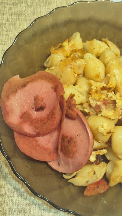 The image shows a close-up view of a plate of food. The plate is dark brown and has a wavy rim. The food consists of pasta, scrambled eggs with bits of what appears to be bacon or ham, and slices of pink sausage. The pasta is shell-shaped and mixed with the scrambled eggs and meat. Two large slices of sausage are prominently placed in the foreground, overlapping each other. The sausage has a slightly fried appearance with some darker brown edges. The overall presentation suggests a home-cooked meal. The background appears to be a light-colored, textured surface, possibly a table. There are no people or specific environmental details that indicate the time of day, weather, or a precise location within Ialoveni, Moldova, beyond what is provided. The lighting suggests indoor illumination. No visible text is present in the image.