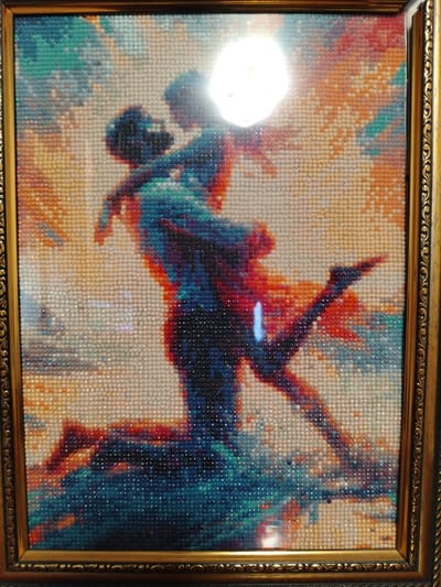 This image is a diamond painting of a couple embracing. The couple is silhouetted against a bright, circular light source, possibly the moon or sun. The woman is being lifted into the air by the man, with her legs extended. They are dressed in flowing garments, and the colors of the painting are vibrant and varied, ranging from deep blues and reds to lighter oranges and yellows. The painting is framed by a decorative gold frame. The location in Geologorazvedka, Russia is not discernible from the image itself.