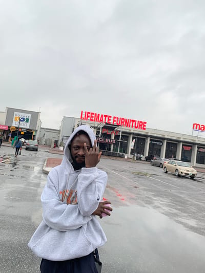 The image captures a person in a gray hoodie standing outside a shopping complex in Sangotedo, Nigeria. The person is facing the camera, holding a pink object in one hand, and making a hand gesture. The shopping complex houses businesses like "LIFEMATE FURNITURE," "UCLEUS RESTAURANT & LOUNGE," "NOVARE," "LEKKI," "BURGER NATION," and "DODO PIZZA." Cars are parked in the complex's parking area, which appears wet from rain. The sky is overcast, suggesting recent or ongoing rain. Other pedestrians are visible in the background near the shopping complex entrance.