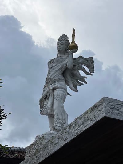 A low-angle shot depicts a white, intricately carved statue of a deity or celestial being, potentially female, against a backdrop of cloudy sky. The statue is positioned as if floating or flying, holding an ornate golden vessel. The deity wears a crown and a flowing garment with decorative elements. A portion of a building structure, possibly part of a temple or other architectural structure, appears in the upper right corner of the image, featuring carved details and dark wooden elements. The location is Celuk, Indonesia, based on the architectural style and cultural context suggested by the sculpture. Vegetation is visible in the lower portion of the frame.