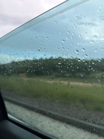 The image shows a view through a wet window, likely a car or train window, in Belo Pole, Bulgaria. Droplets of rain obscure the background, which appears to be a combination of a cloudy sky and a blurred landscape of trees and a road or railway. A portion of the window frame is visible in the foreground, and a dark interior element is present at the bottom right corner.