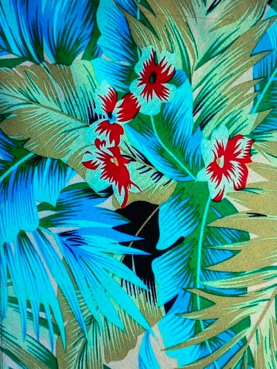 The image showcases a vibrant tropical pattern. Predominantly, palm-like leaves in shades of turquoise, green, and gold interweave, creating a dense foliage effect. Striking red flowers with white outlines are scattered throughout, adding a pop of color. A dark patch in the center disrupts the continuity of the pattern, possibly indicating a shadow or negative space. The overall composition presents a lively and colorful depiction of a tropical motif.