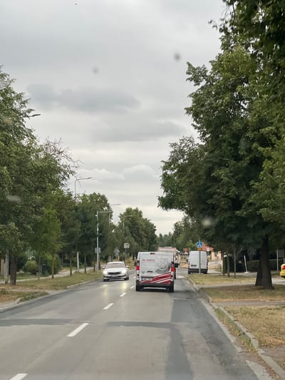 The image shows a street scene in Měnín, Czechia. The weather is overcast. On the right side of the road, a row of green trees lines the sidewalk. A white van with red and white decals that reads "ML PARTS" is driving in the middle lane, followed by another white van. A white sedan is in the left lane. Further down the road, there are more trees and buildings. The road has white lane markings. There are streetlights visible along the road.