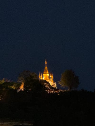 A night time photograph captures the illuminated golden spires of Wat Xieng Thong in Luang Prabang, Laos. The temple's distinctive tiered roofs and finials are brightly lit against a dark, almost black, night sky. Silhouetted trees in the foreground partially obscure the base of the temple structure, creating a sense of depth and framing the sacred site. The overall impression is one of quiet reverence and architectural beauty in the darkness.