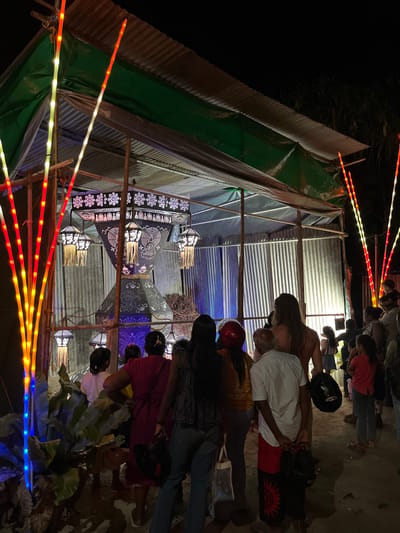 The image depicts an outdoor scene at night, likely in Habaraduwa, Sri Lanka. A group of people are gathered, observing or participating in an event centered around illuminated lanterns. The lanterns are intricately designed with decorative patterns, some appearing to be made of paper or fabric stretched over frames, and are lit from within, casting a warm glow. They hang from a makeshift structure supported by bamboo poles, which also appears to have corrugated metal roofing and green tarpaulin sides, suggesting a temporary setup, possibly for a festival or market stall.
Adding to the festive atmosphere, colorful LED string lights in red, yellow, and blue hues are strung along the foreground and sides of the structure, creating a vibrant and celebratory ambiance. The time of day is clearly night, evidenced by the darkness and the reliance on artificial lighting. The ground appears to be sandy or dusty. Some individuals are wearing helmets, possibly indicating they arrived on motorcycles. The overall mood seems to be one of community gathering and enjoyment, with the focus on the decorative lights and lanterns. No visible text is present in the image.