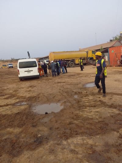A group of people are gathered on a muddy construction site near a white van and a large yellow tanker truck. The ground is covered in puddles and dirt. A man wearing a yellow hard hat and work clothes walks in the foreground. Behind him, the group stands clustered together, possibly in discussion. The sky is overcast, adding to the drab atmosphere of the scene. A building and shipping container are visible in the background.