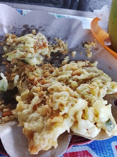 The image shows a close-up of a plate of fried food, possibly tempeh mendoan, a popular Indonesian snack. The fried items are golden brown and crispy, with visible pieces of tempeh and some vegetables like carrots and green onions. They are served on a piece of brown paper with a scalloped edge, which rests on a blue and red checkered tablecloth. A small orange dish with a white napkin and part of a green bottle or glass is visible in the upper right corner. The lighting suggests an outdoor setting, possibly at a street food stall or a casual eatery in Adipala, Indonesia. The focus is on the food itself, with the background blurred.