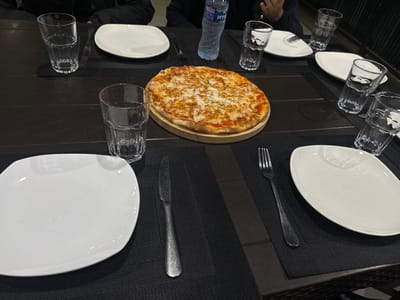 A pizza sits on a wooden board at the center of a dark-colored table. Several white plates are positioned around the table, each accompanied by a glass. Some plates also have a fork and a knife beside them. A water bottle is placed at the head of the table. The setting suggests a group meal is about to be enjoyed.