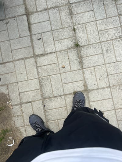 An overhead, ground-level shot captures a person's lower body, focusing on their legs and feet, on a light grey paved surface in Beloslav, Bulgaria. The pavement is composed of rectangular concrete blocks laid in an interlocking pattern. The person wears black pants, a partially visible white shirt, and dark grey and black sneakers with prominent lacing and a textured upper design. Small, partially visible text, possibly "SL", is visible on the side of the left shoe.
The paved area is strewn with minor debris, including dried grass blades, a small light-brown pebble, and a dark cylindrical object (possibly a cigarette butt). Patches of dirt, some green weeds, and a crumpled white item are also visible along the edges and between the blocks. The lighting appears even and diffused, suggesting daytime with possible overcast conditions. A small, unreadable white text or logo is present on the sleeve of the black garment worn by the person. The image provides a close-up perspective of the ground and the footwear.