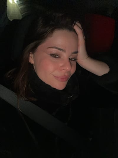 A woman with fair skin and brown hair poses inside a vehicle at night. She rests her hand on her head and smiles slightly. A seatbelt crosses her chest. She wears a black turtleneck sweater. The lighting is dim, with some light reflecting on her face and illuminating a red headrest in the background.