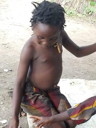 The media file shows a young child, likely a boy given the clothing and general appearance, sitting outdoors in what appears to be a rural setting in Otulu, Nigeria. The child has dark skin and dreadlocks, and is wearing a patterned cloth around their waist, with red, yellow, and black geometric designs. Around the child's neck, a gold-colored crucifix necklace is visible.
The child is looking down, and their facial expression is somewhat obscured, but the posture suggests a contemplative or perhaps slightly sad mood. The background shows a dirt ground and some dry vegetation or reeds. The lighting suggests it is daytime, possibly with overcast conditions as there are no strong shadows. Another person's arm, wearing a similar patterned fabric, is visible in the foreground, reaching towards the child, suggesting an interaction or caretaking presence. The overall scene depicts a simple, everyday moment for a child in this location. No visible text is present in the media file.