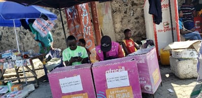 The scene depicts a street vendor selling mobile phone accessories, located at coordinates -1.668 latitude, 29.268 longitude in Rubavu, Rwanda. Two individuals are seated behind pink "Africell" kiosks with advertising signage. One man is dressed in green, looking down at a mobile phone. The other man is wearing a black hat and a pink vest, facing forward. Another man, dressed in red, is seated at a nearby, partially obscured kiosk. A fourth man, dressed in blue, is standing next to a blue umbrella, observing the scene.  A table filled with miscellaneous items is located on the left side of the scene. There are boxes and plastic containers behind the kiosks. Graffiti is visible on the wall behind the vendors. The scene is lit by natural sunlight and appears to be in a street market setting.