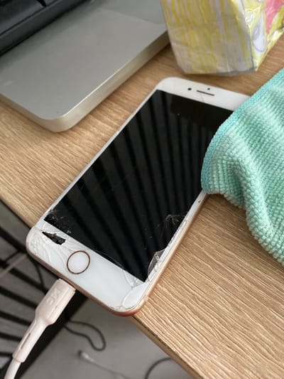 The image shows a close-up of a damaged smartphone placed on a wooden desk. The phone's screen is cracked, and the top right corner of the phone is visibly broken, with pieces missing. A white charging cable is plugged into the bottom of the phone. To the right of the phone, a teal-colored microfiber cloth is partially visible, and behind that, a colorful, paper-wrapped object, possibly a gift or toy, is present. In the background, the edge of a silver laptop is visible. The overall scene suggests a casual indoor setting, possibly a home or office. The location of Maksimkovo, Russia, is not depicted in the image and cannot be inferred from its content.