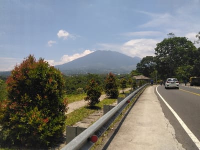 The image depicts a roadside scene in Cugenang, Indonesia, under clear daylight conditions. In the immediate foreground to the left, a dense, round-shaped bush with prominent green and reddish foliage is present. A silver-colored metal guardrail with red circular reflectors extends from the lower left to the mid-center, running parallel to a concrete pedestrian path. Grass and smaller ornamental bushes line the area between the guardrail and the path.
To the right of the guardrail and path is an asphalt road, marked with a solid white line on the shoulder and a broken white lane divider further inward. A white sedan is visible traveling away from the camera in the right lane, and the front portion of a yellow utility truck is partially visible on the far right shoulder. Several indistinct human figures are dispersed near the roadside further down, with one individual appearing to walk or cycle near a small, open-sided shelter with a light-colored roof, positioned behind the guardrail and bushes.
The background is dominated by a large, forested mountain, likely Mount Gede or Pangrango, partially obscured by atmospheric haze. Below the mountain, a sprawling urban or semi-urban settlement is visible within a valley. The sky above is clear blue with scattered white cumulus clouds. Tall green trees line the right side of the road, contributing to the lush environment.