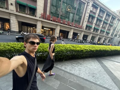 The image, oriented correctly, depicts an urban street scene during daytime. In the foreground, a male individual is taking a selfie, holding the camera (implied) with an outstretched arm. He is wearing a black sleeveless top and black sunglasses, with short brown hair. His expression is neutral due to the sunglasses. Behind him, slightly to the left, a female individual walks away from the camera. She wears a black dress or skirt, purple sandals, and sunglasses, with her light brown hair pulled back.

The subjects are on a paved sidewalk, which features various patterns of gray pavers. A low, neatly trimmed green hedge separates the sidewalk from a street. Along the street, a row of parked motorcycles and scooters is visible. A dark-colored car can also be seen.

Across the street stands a large, multi-story building, characteristic of modern urban architecture, with numerous windows and balconies featuring green awnings. The ground floor of the building appears to house retail establishments.

The lighting is bright, suggesting daylight, possibly overcast or late afternoon, as shadows are not sharp. The sky, visible above the building on the left, appears clear.

Prominently displayed on the building across the street are large red Chinese characters that read "天幕MALL" (Tiān mù MALL), translating to "Sky Curtain Mall." Below this text, a shield-shaped logo is visible on the building facade. Faintly visible in one of the ground-floor windows is the brand name "Nikon." The overall setting suggests a bustling commercial area within an East Asian city, consistent with the provided context of Putuo District, China.