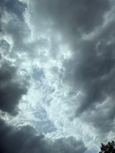 The image displays a dramatic sky filled with dark, heavy storm clouds interspersed with patches of brighter, lighter clouds. A sliver of blue sky is visible through a gap in the clouds, suggesting the storm is either approaching or moving away. The lighting is dim and diffused, typical of pre-storm or stormy conditions. The bottom right corner of the image shows the dark green foliage of a tree, indicating an outdoor setting, possibly on the outskirts of a populated area. There are no people or specific man-made structures visible in the frame. The overall impression is one of impending or ongoing inclement weather. The location in the city of Аур, Russia, is not identifiable from the visual information present in the image itself.