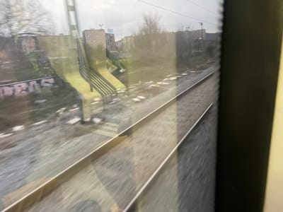 A view from inside a moving train reveals a gray, urban landscape. The railroad tracks stretch into the distance, with gravel and debris scattered around. Buildings are visible through the window, including graffiti-covered walls and high-rise apartments. There is reflection of a yellow escalator on the window of the train. The sky is overcast, adding to the somber feel of the scene.