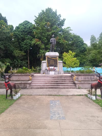 The image shows a statue of a man on a pedestal in Baan Thong Nang, Thailand. The statue stands atop a series of steps leading up to a concrete structure. The pedestal features a plaque with text in Thai. In front of the steps are two deer statues, each adorned with a red floral garland. The ground is paved, with greenery visible to the sides. Behind the structure are numerous trees forming a dense backdrop. The sky is overcast.