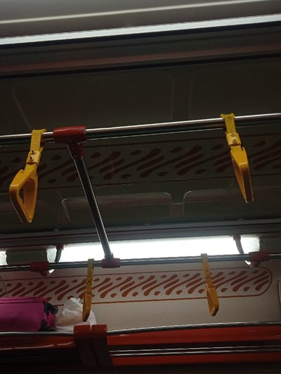 The image shows the interior of a bus, likely in the area of Kecamatan Prambanan, Indonesia, based on the coordinates provided. The image captures the ceiling of the bus, showcasing two yellow handrails, one of which is connected to a silver, extending handrail that is secured by a red bracket. The ceiling is adorned with a decorative pattern featuring red wavy lines on a white background. A fluorescent light is illuminating the scene. Beneath the handrails, a white shelf is visible, with a pink bag and a white plastic bag resting on it. Another red bracket is visible, and the image captures the underside of an overhead red bar. There are no people visible in the image. The scene appears to be a typical bus interior.