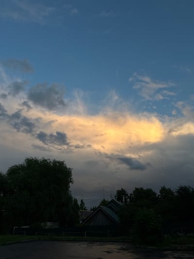 The image depicts a cloudy sky with a striking band of golden-yellow illuminated clouds stretching horizontally across the frame. Below the sky, the silhouettes of trees and rooftops are visible against the darkening landscape. In the foreground, a wet asphalt surface suggests recent rainfall. The scene appears to be a rural or suburban setting, possibly within the city of Zakharovo, Russia, given the architectural style of the buildings and the dense tree cover. There are no people or overt actions visible in the image. The overall mood is somber and atmospheric, with the dramatic lighting of the clouds contrasting with the shadowed foreground.