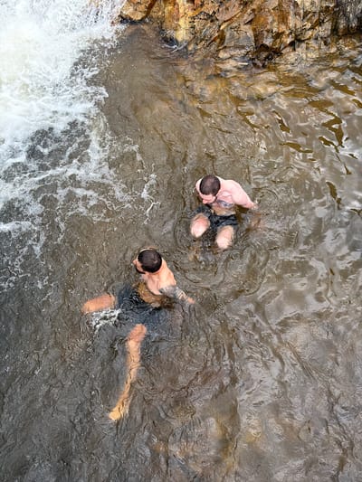 Two men are standing in shallow water near a rocky embankment and a small waterfall. The water is murky brown and has some white foam on the surface. The man in the foreground is facing away from the camera, looking towards the waterfall. He has dark hair and is wearing black swim shorts. The man in the background is also facing away from the camera, his head and shoulders visible above the water. He has short dark hair and is wearing dark swim shorts. Both men appear to be of similar build. The rocks behind them are varied in color, with shades of brown, orange, and gray. The waterfall is on the left side of the image, with white water cascading down. The overall scene suggests a natural outdoor setting, possibly a river or creek.