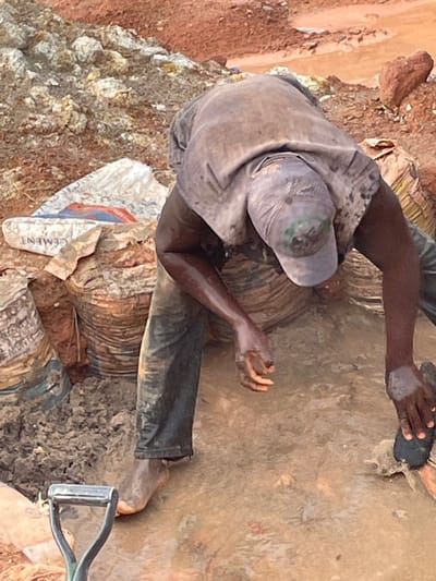 The image depicts two individuals engaged in manual labor in a muddy, outdoor environment, likely a construction or earth-moving site in Rayfield, Nigeria. The person in the foreground, viewed from behind and slightly to the side, is wearing a dirty grey t-shirt, dark pants, and a grey baseball cap. Their hands are visibly caked with mud. They appear to be sifting or processing some material.

The second person, partially visible in the upper right of the frame, is also hands-on, with mud on their arms and hands. They are holding a dark, flat object, possibly a tool or material being manipulated. The ground is a mixture of soil, mud, and what appears to be aggregate or debris. A torn white bag, with visible red and blue lettering and the word "CEMENT" printed on it, lies on the ground near the foreground subject, suggesting construction-related work. A shovel handle is visible in the lower right corner, further reinforcing the context of manual labor involving earth and materials. The lighting suggests it is daytime, with no clear indicators of specific weather conditions beyond the presence of mud. The overall impression is one of hard work in challenging conditions.