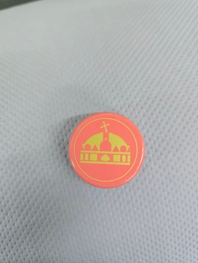 A close-up, slightly high-angle shot shows a round, coral-pink pin with a yellow outline of a crown featuring a cross on top. The pin rests on a textured, light blue surface. The image does not provide any information about the city of Worms, Germany, or any people, actions, or interactions.
