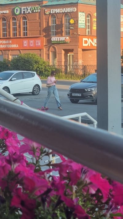 A street scene in the city of Megion, Russia. In the background is a brick building with businesses labeled "FIX PRICE," "ЕРМОЛИНО," "ОПТИКА," and "DNS." In the foreground, a woman wearing a pink shirt and blue jeans walks across the street, looking at her phone. A white car is parked on the left side of the street, and a gray car is driving on the right. Pink flowers are in the foreground, obscuring the bottom of the image.