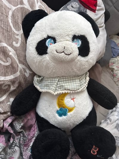A plush panda toy is positioned in the foreground, lying on a bed. The panda is white with black ears, eye patches, arms, and legs. It wears a light green and white checkered collar and has a crescent moon and cloud embroidery on its belly. The background consists of bedding with a patterned duvet cover, featuring floral and abstract designs in brown, white, and pink. The scene is indoors, likely in a bedroom. The specific location in the city of Wołomin, Poland, cannot be determined from the image.