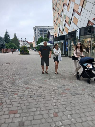 In Truskavets, Ukraine, a cloudy day unfolds on a cobblestone street. A man in a black t-shirt and shorts stands with his hands on his hips, looking to his left. Beside him, a woman with dark hair, wearing a striped shirt and black skirt, walks away from the camera, carrying a white bag. Further down the street, another woman in a light pink sweater pushes a stroller with a baby inside. The background features modern buildings with geometric facades and glimpses of shops. Green trees and bushes line the street, suggesting a well-maintained urban park or pedestrian area. The overall atmosphere appears to be a casual stroll on a day with overcast skies. There is no visible text in the image.