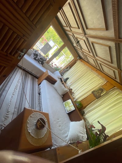The interior of a room, situated in General Todorov, Bulgaria, is observed from an elevated perspective, looking downwards. The room features a prominent wooden aesthetic with a decorative coffered ceiling composed of rectangular panels and ornate wooden molding. A single bare light bulb is suspended from the ceiling.
A large window or door assembly, occupying the upper left section of the room, offers a view of a bright, sunny exterior. Green foliage and red blossoms are visible outside, along with two freestanding informational signs, one of which displays a portrait of an individual and text.
The floor is covered by a striped rug featuring white and dark grey/brown vertical stripes, accented with fringed edges. A substantial light-colored rope, with darker flecks, extends diagonally across the room, originating near the window and descending towards the lower right.
Two beds or daybeds are arranged within the space. One, on the right side of the room, is covered with a white, intricately crocheted bedspread and two matching pillows. The other bed or bench, partially visible on the left, also features a white covering with a fringed edge.
Along the window and wall areas, multiple wooden ledges or shelves are utilized for display. Objects on these ledges include: a black and white photograph depicting a group of people, a small framed mirror reflecting outdoor greenery, a potted plant, a ceramic dog figurine, a small white dome security camera mounted on a wooden beam, and an Orthodox icon portraying the Virgin Mary and Child. Below parts of these ledges are pleated, cream-colored curtains.
In the foreground, a low, dark wooden table or bench supports a white crocheted doily, on which rests a dark bowl containing several metallic coins. Near one of the beds, on the floor, a white-framed picture is visible, alongside a vase holding dark red/purple calla lilies.
No individuals are physically present within the room; however, human figures are depicted in the photograph and the icon. The room is brightly illuminated by natural light entering through the window, indicating a daytime scene.