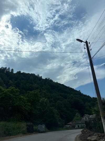 The image shows a rural road in Gorno Trape, Bulgaria. The road curves to the right, bordered by lush green trees and vegetation on the left. Several houses with tiled roofs are visible nestled amongst the trees on the hillside. A dark, wooded hill dominates the background. A utility pole with a street lamp and overhead wires stands prominently on the right side of the road. Two large trash bins are situated on the left side of the road, near the edge of the vegetation. The sky above is partly cloudy with a mix of white and grey clouds, and patches of blue sky.