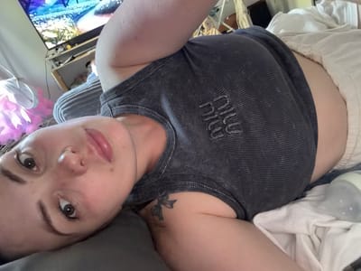 A young woman with brown eyes, a small nose ring, and full lips lies on her side on a bed. She is wearing a dark gray tank top with a ribbed texture and white lettering that appears to say "MIU MIU". A small bow tattoo is visible on her left shoulder. Her light-colored shorts are visible just above the waistband of her tank top. A white towel and a baby bottle with white liquid inside are next to her on the bed. In the background, a television screen displays a colorful image. The overall scene appears to be a bedroom.