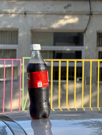 A Coca-Cola Zero Sugar bottle, 500 ml, stands on a dark, reflective surface, likely the hood of a car, in an outdoor setting. The bottle is condensation-covered and partially filled with a dark liquid. The label is visible, showing text in Cyrillic script, including "Coke БЕЗ ЗАХАР" (Coke WITHOUT SUGAR) and nutritional information. In the background, a building with a light-colored facade and visible windows is present. A brightly colored railing, featuring pink, yellow, and green bars, runs horizontally across the mid-ground. The image appears to have been taken in Kaspichan, Bulgaria, given the Cyrillic text on the bottle, indicating a specific location within the city. No people are present in the frame.