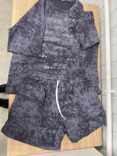The image displays a two-piece clothing set laid out on a light wooden table. The set consists of a t-shirt and shorts, both in a dark gray color with a distressed, textured pattern. The t-shirt features the word "shine" repeatedly printed across it in a lighter, possibly glittery font. The shorts have a drawstring waist and the same textured pattern. The background shows a white wall with horizontal paneling and a white pipe running across the top. There are also hints of what appear to be feet wearing dark shoes at the bottom of the image, suggesting a person is present but not fully visible. The scene appears to be an indoor setting, possibly a room in a home or a retail space, given the simple furnishings. There is no indication of time of day or weather conditions. The overall impression is of clothing being presented or displayed. The visible text is "shine" repeated on the t-shirt.