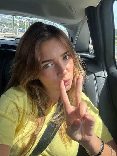 The image captures a young woman inside a car, likely in Torres Vedras, Portugal, taking a selfie or being photographed from an elevated angle. She lies back with her head tilted, looking directly at the camera with a relaxed expression and slightly pursed lips. Her long, brown hair, with visible lighter streaks, cascades around her face and over her bright yellow long-sleeved top or sweater.
Her right hand is raised, forming a "peace sign" or "victory sign" with her index and middle fingers extended. On her right wrist, she wears multiple bracelets, including a silver-toned chain and a black-and-silver link design. A ring is visible on her left ring finger. A dark seatbelt diagonally crosses her chest.
The car's interior features dark upholstery and trim. Through the windows, a bright, blurry outdoor scene is visible, suggesting an urban or semi-urban landscape with buildings and utility poles, illuminated by strong sunlight. The bright lighting indicates it is daytime and sunny. No visible text appears in the image.