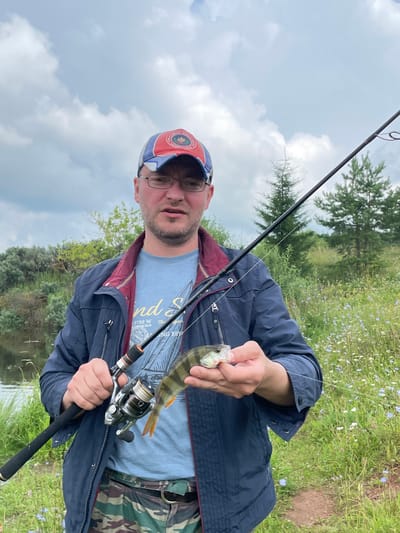 A medium shot captures a man holding a small fish and a fishing rod on a cloudy day. The man, wearing glasses and a baseball cap, faces the camera with an open mouth, suggesting he is speaking. He is dressed in a blue jacket over a t-shirt and camouflage pants. The fishing rod, with a reel, extends from the right side of the frame. The background features a body of water on the left and a field of wildflowers and trees on the right, indicating a rural or natural setting. The specific location, the city of Кумёны, Russia, places this scene within a region known for its natural landscapes and fishing opportunities.