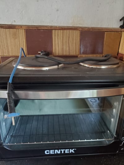 The image shows a black countertop oven with a glass door, branded "CENTEK". The oven is positioned on a counter against a tiled backsplash. A blue electrical wire, taped to a metal object, extends from the top of the oven and drapes over the side. The interior of the oven contains a wire rack. The scene is lit by ambient light. The location is identified as Konstantinovka, Moldova.