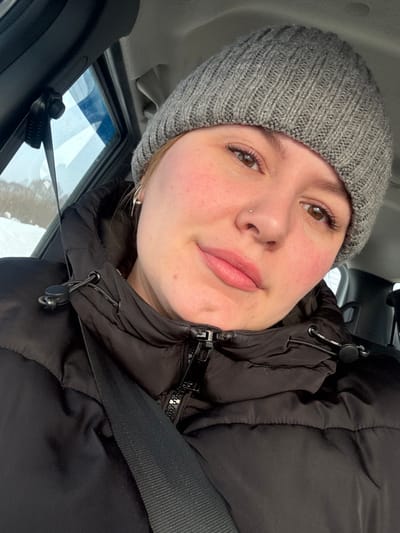The scene depicts a female individual inside a vehicle. As per the query, the location is the city of Laiga, Russia, though no specific geographical identifiers are visible in the image itself. The individual, appearing to be an adult, is oriented towards the camera, displaying a neutral expression. She wears a grey knitted beanie and a black puffer jacket with a visible front zipper and drawstring collar details. A black vehicle seatbelt is positioned diagonally across her chest and left shoulder. Her visible facial features include light skin with areas of redness on the cheeks, brown eyes with distinct eyelashes, and pink-toned lips. A small, light-colored stud piercing is present on her left nostril, and a silver-colored hoop earring is visible on her left ear. Through the vehicle's left-side window, a blurred outdoor environment is visible, characterized by snow-covered ground and what appears to be bare vegetation, suggesting a winter setting.