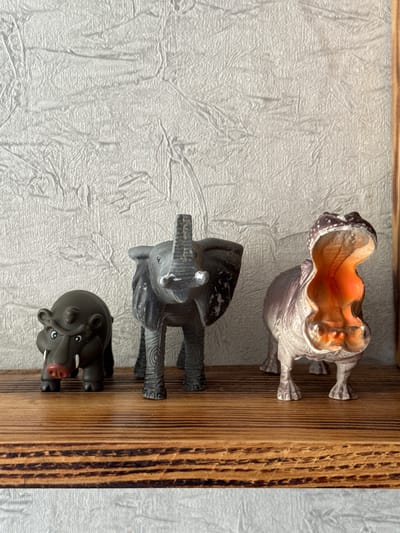 A close-up, eye-level shot shows three toy animals lined up on a wooden shelf against a textured, light-colored wall. The animals, from left to right, are a small, dark gray hippo with a red nose, an elephant with its trunk raised, and a large, light brown and purple hippopotamus with its mouth wide open revealing an orange interior. The shelf has a visible wood grain pattern. The background wall has a rough, stucco-like texture. This scene is located in the Яромінскі сельскі Савет, Belarus.