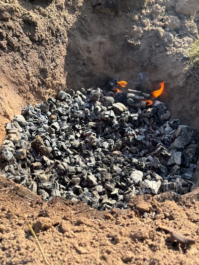 An outdoor scene shows a pit dug into the earth, filled with smoldering charcoal. Small, orange flames flicker within the charcoal, producing wisps of smoke. The edges of the pit are formed by soil, with some dry grass visible at the top right. The lighting suggests a sunny day. The overall impression is of a fire pit in an outdoor, possibly rural, setting. There are no people visible in the image.